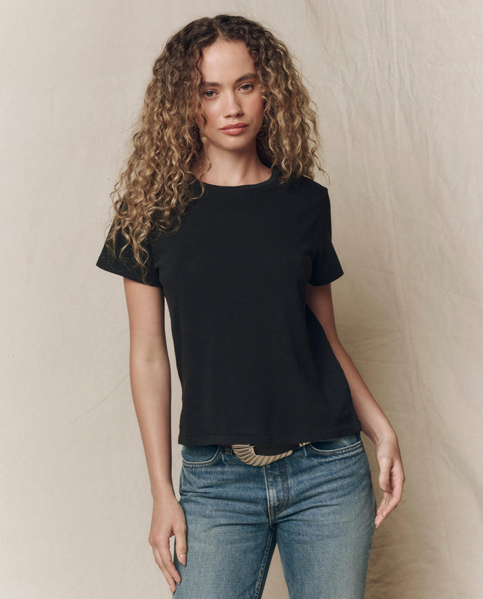 A person with curly hair wears THE GREAT. SP23 CORE KNITS The Little Tee. Solid -- Almost Black and blue jeans, standing against a neutral backdrop—an ideal layering tee for effortless style.