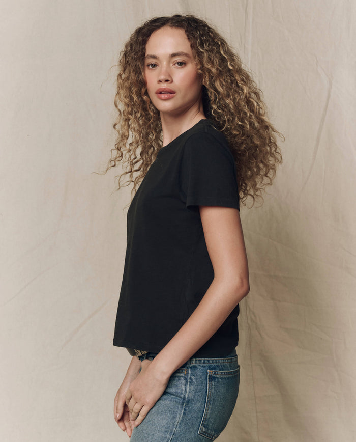 A person with curly hair wears THE GREAT. SP23 CORE KNITS The Little Tee. Solid -- Almost Black and blue jeans, standing sideways against a neutral fabric backdrop.