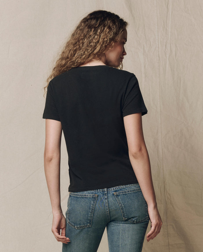 A person with curly hair wears THE GREAT. SP23 CORE KNITS The Little Tee. Solid -- Almost Black and blue jeans, standing with their back to the camera against a neutral background.