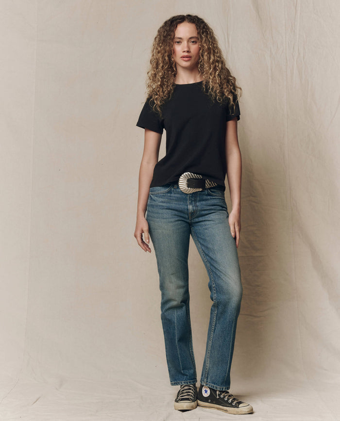 A person with curly hair stands against a beige backdrop, wearing THE GREAT. SP23 CORE KNITS The Little Tee. Solid -- Almost Black, blue jeans with a large belt buckle, and black Converse sneakers.