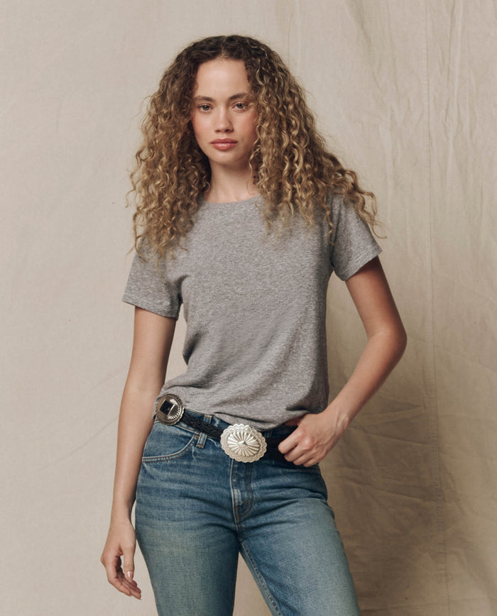 A person with curly hair wears THE GREAT.'s The Little Tee. Solid in Heather Grey (SP22 APRILCAP), blue jeans, and a decorative belt with a large silver buckle, standing against a neutral fabric backdrop.