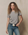 A person with curly hair wears THE GREAT.'s The Little Tee. Solid in Heather Grey (SP22 APRILCAP), blue jeans, and a decorative belt with a large silver buckle, standing against a neutral fabric backdrop.