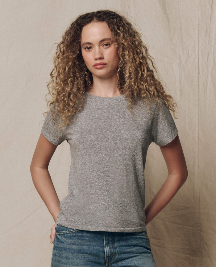 A person with curly hair wears THE GREAT. SP22 APRILCAP's The Little Tee. Solid in Heather Grey and blue jeans, standing before a neutral fabric backdrop.