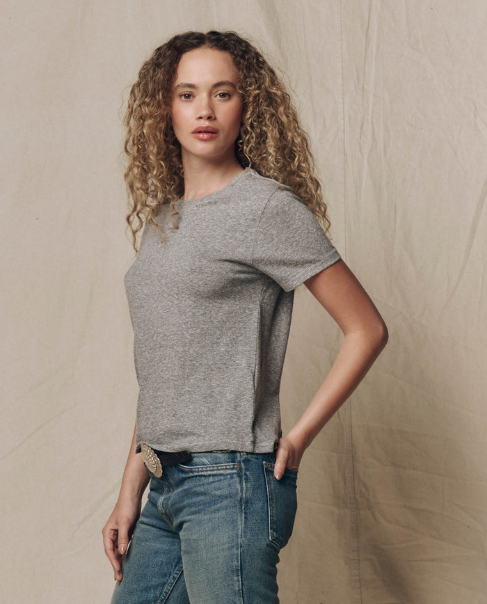 A person with curly hair wearing THE GREAT. SP22 APRILCAP's The Little Tee in Heather Grey stands before a beige fabric backdrop, gazing at the camera.