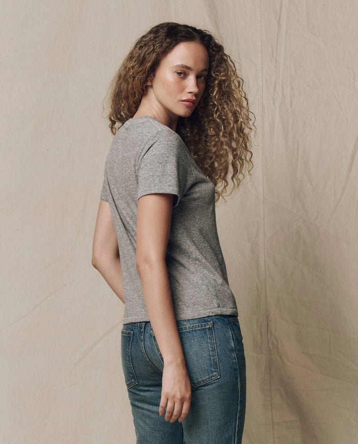A person with curly hair stands sideways, looking at the camera against a neutral fabric backdrop, wearing THE GREAT. SP22 APRILCAP The Little Tee. Solid in Heather Grey and blue jeans.