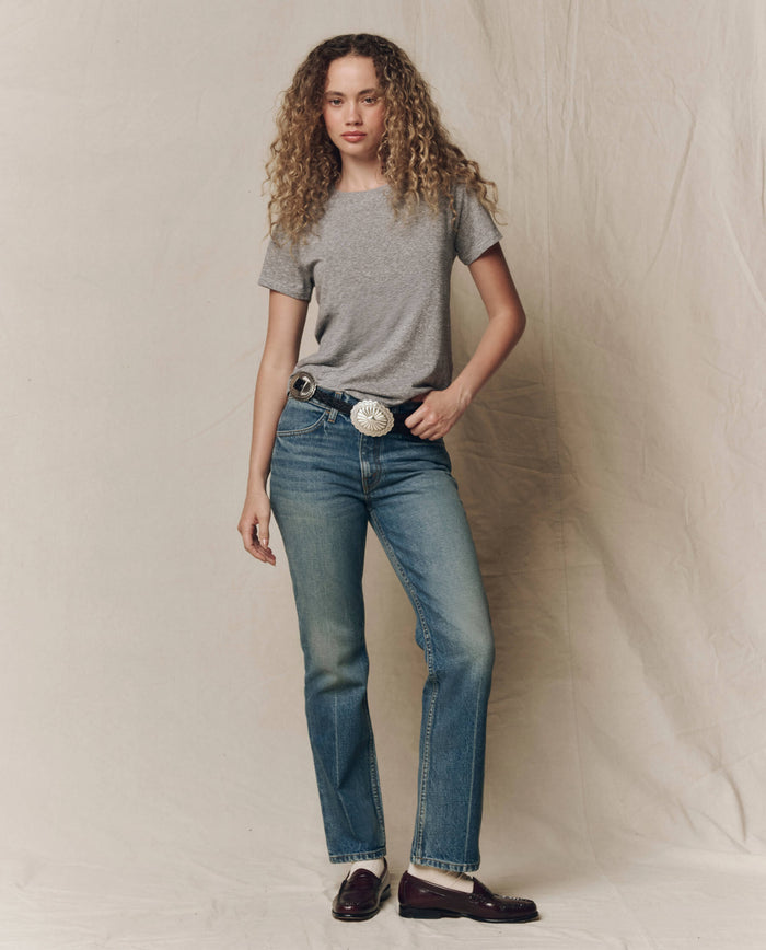 A woman with curly hair stands against a plain backdrop, wearing The Little Tee. Solid in Heather Grey by THE GREAT. SP22 APRILCAP as a layering piece, paired with blue jeans, a large belt, and dark loafers.