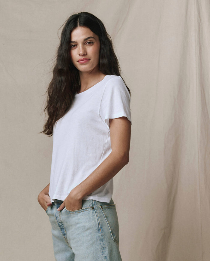 A woman with long dark hair stands with hands in pockets against a neutral fabric backdrop, wearing THE GREAT. SP22 APRILCAP's The Little Tee in True White and light blue jeans.