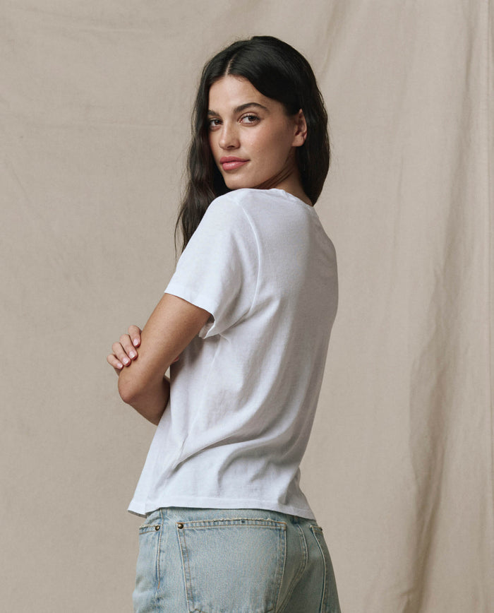 A woman with long dark hair stands sideways, arms crossed, wearing THE GREAT. SP22 APRILCAP The Little Tee. in True White and light blue jeans against a neutral beige background.