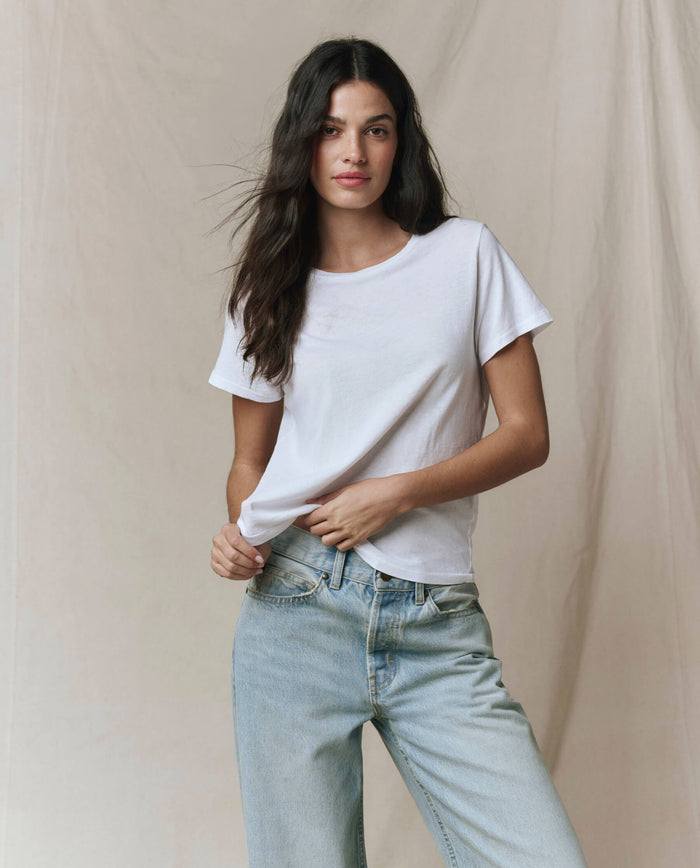 A woman with long dark hair wears THE GREAT.'s The Little Tee in True White and light blue jeans, standing against a beige fabric backdrop.
