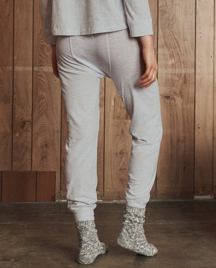 A person wearing THE GREAT. H25 SLEEP The Long John in Heather Grey stands on a wooden floor in front of a wood panel wall.