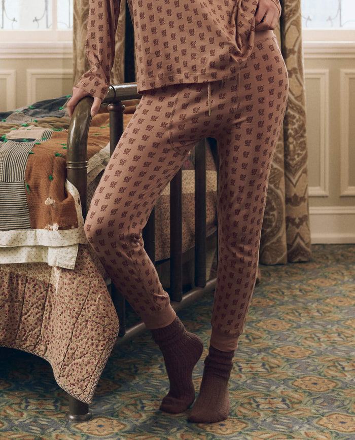 A person wearing matching THE GREAT. F26 SLEEP The Long John. -- Rose Print pajamas and socks stands next to a vintage metal bed in a carpeted room with patterned curtains and bedding.