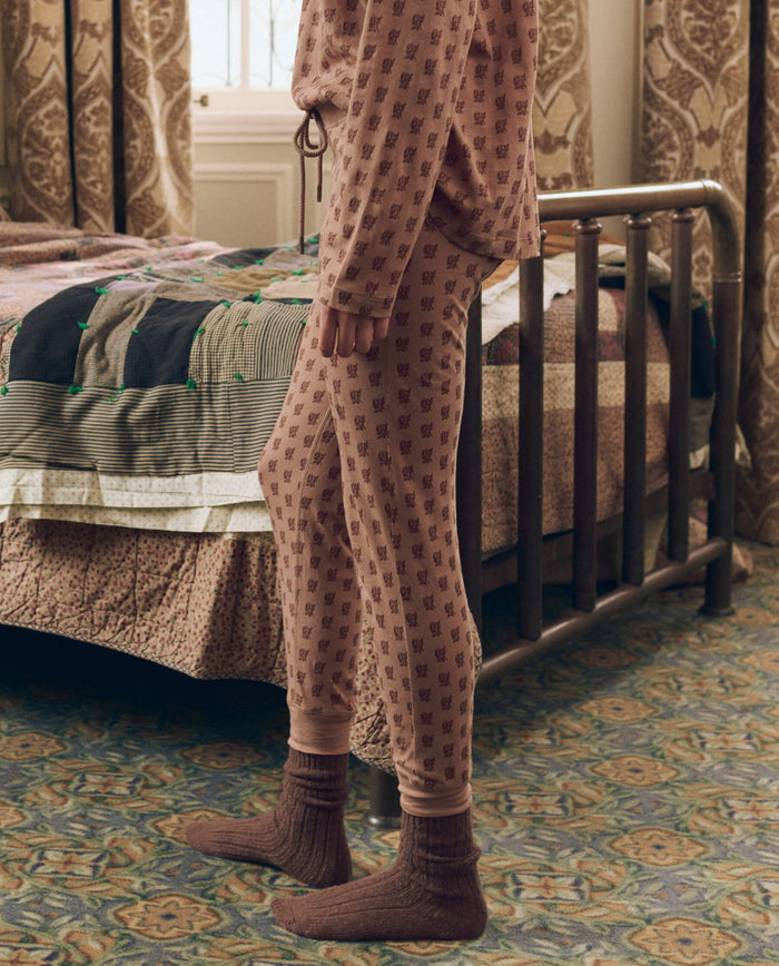Wearing The Long John in Rose Print from THE GREAT. F26 SLEEP, a person with socks stands beside a made bed in a carpeted bedroom with patterned curtains.