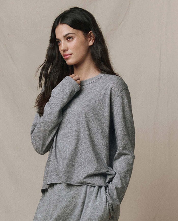 A woman with long dark hair wears THE GREAT. CORE KNITS The Long Sleeve Crop Tee in Heather Grey, paired with matching pants, standing against a neutral background.