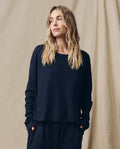 A woman with long blonde hair, wearing The Long Sleeve Crop Tee in True Navy by THE GREAT. FALL 23 KNITS along with dark pants, stands against a beige backdrop, looking to her right.