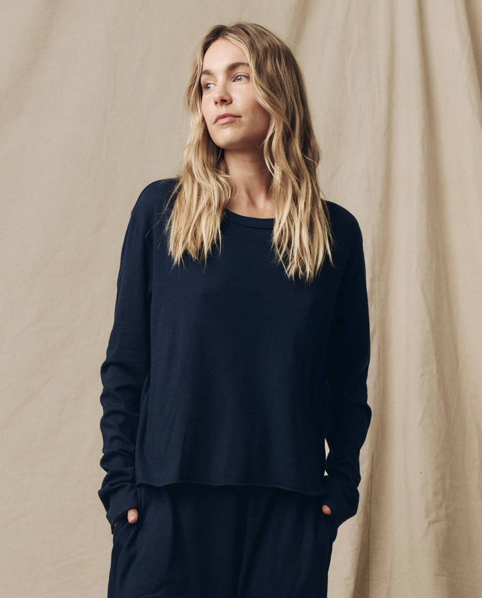 A woman with long blonde hair, wearing The Long Sleeve Crop Tee in True Navy by THE GREAT. FALL 23 KNITS along with dark pants, stands against a beige backdrop, looking to her right.