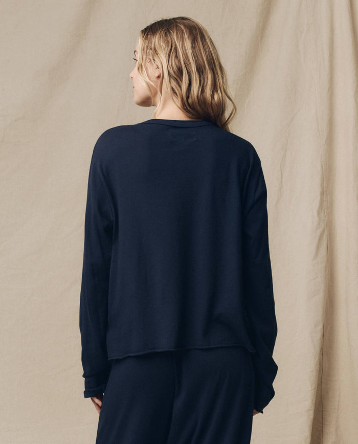 A person with long hair wearing THE GREAT. FALL 23 KNITS' The Long Sleeve Crop Tee in True Navy stands with their back to the camera against a neutral backdrop.
