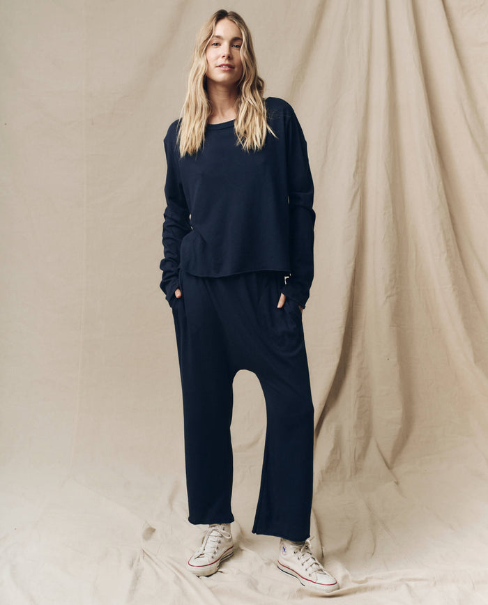 A person stands against a beige backdrop, donning THE GREAT.'s Fall 23 "The Long Sleeve Crop Tee" in True Navy—crafted from 100% cotton. The outfit features loose-fitting long sleeves and pants, complemented by white sneakers, with their hands tucked into their pockets.