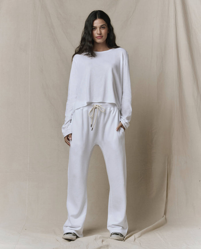 A person stands against a beige fabric backdrop, wearing THE GREAT. CORE KNITS' The Long Sleeve Crop Tee in True White with raw hem details, styled with matching drawstring pants and sneakers.