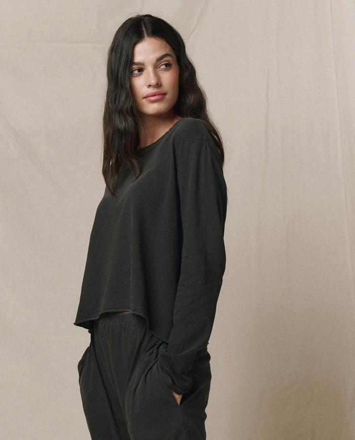 Woman with long dark hair wears THE GREAT. CORE KNITS The Long Sleeve Crop Tee in Washed Black, paired with matching black pants, standing with hands in pockets against a neutral background.