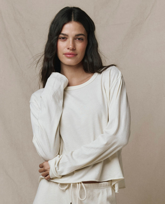 A woman with long dark hair wears THE GREAT. CORE KNITS' The Long Sleeve Crop Tee in Washed White, standing before a plain beige backdrop.