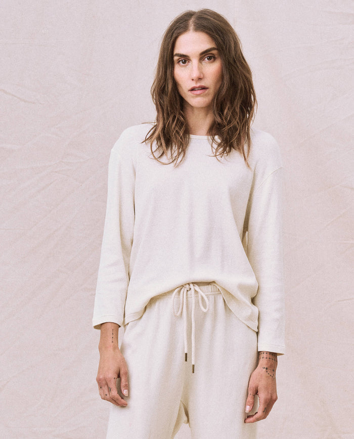 Against a plain background, a person with long hair is wearing THE GREAT. HOL 22 D1's The Long Sleeve Recycled Slim Crew in Natural, crafted from a supersoft and lightweight recycled cotton blend, paired with matching pants.