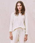 Against a plain background, a person with long hair is wearing THE GREAT. HOL 22 D1's The Long Sleeve Recycled Slim Crew in Natural, crafted from a supersoft and lightweight recycled cotton blend, paired with matching pants.