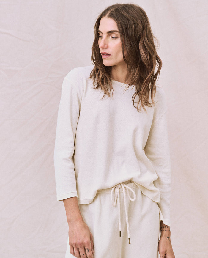 A person with long hair is wearing a light-colored "The Long Sleeve Recycled Slim Crew" top from THE GREAT. HOL 22 D1 and drawstring pants, standing against a neutral background.