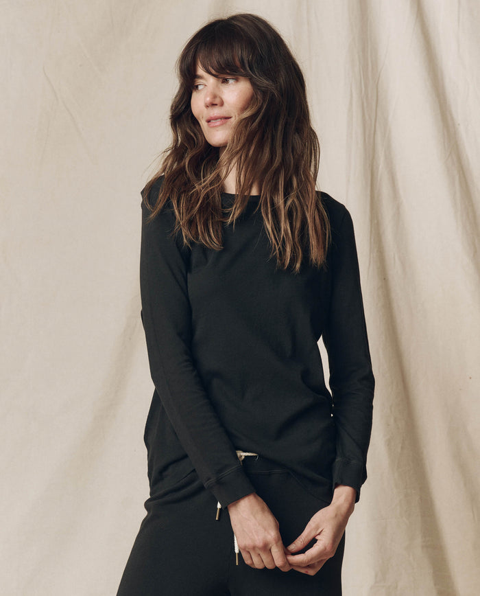 A woman with long brown hair wears THE GREAT. F25 CORE KNITS The Long Sleeve Slim Tee in Solid -- Almost Black, paired with matching 100% cotton pants, as she stands before a beige fabric backdrop.
