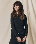A woman with long brown hair wears THE GREAT. F25 CORE KNITS The Long Sleeve Slim Tee in Solid -- Almost Black, paired with matching 100% cotton pants, as she stands before a beige fabric backdrop.