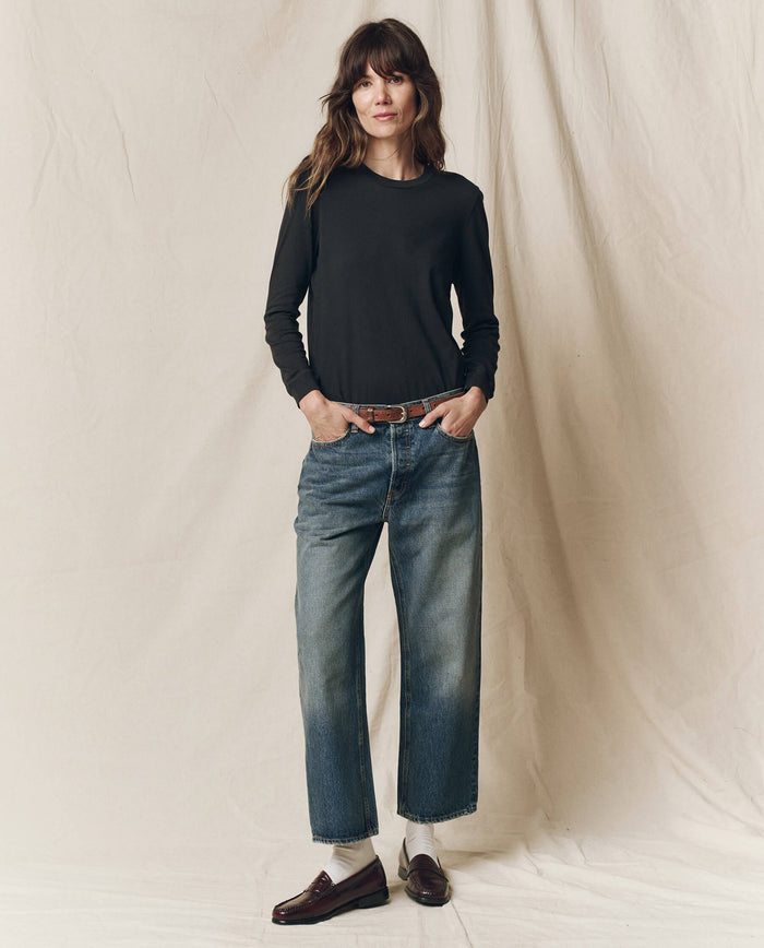 A woman stands against a beige fabric backdrop, wearing THE GREAT. F25 CORE KNITS The Long Sleeve Slim Tee in Solid -- Almost Black, paired with blue jeans, a brown belt, white socks, and brown loafers.