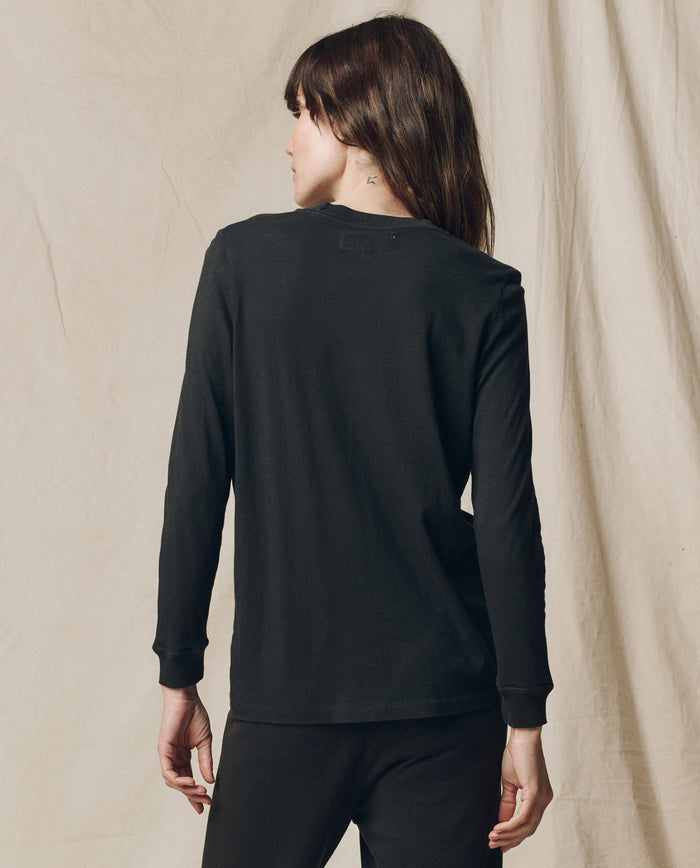 A woman with long brown hair faces away from the camera, wearing THE GREAT. F25 CORE KNITS The Long Sleeve Slim Tee in Solid -- Almost Black, paired with black pants against a neutral fabric backdrop.