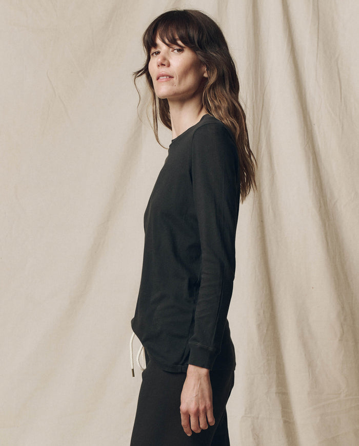 A woman with long brown hair wears THE GREAT. F25 CORE KNITS The Long Sleeve Slim Tee in Solid -- Almost Black and black pants, standing sideways against a beige fabric backdrop. This 100% cotton tee is effortlessly chic.