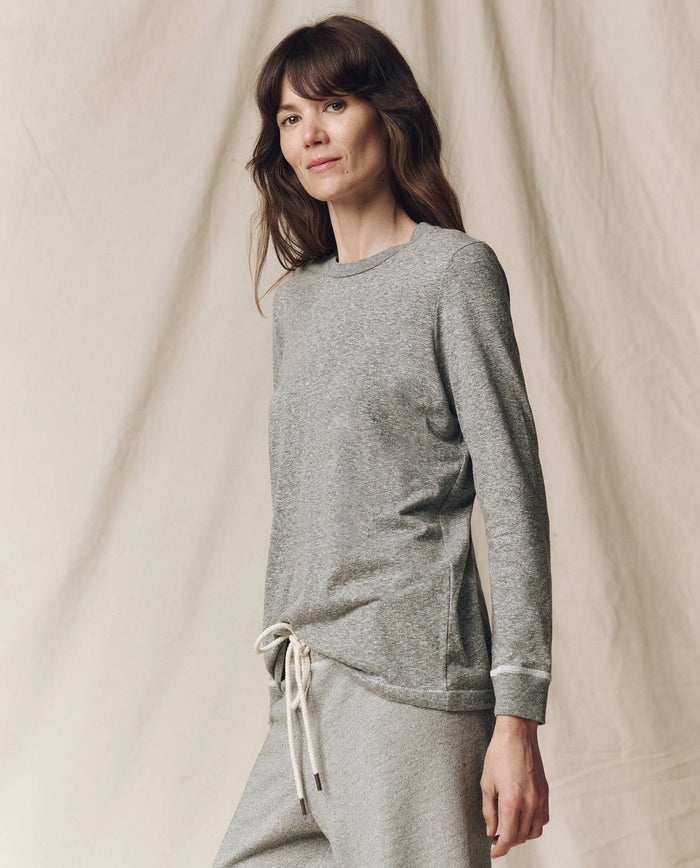 A woman stands before a neutral backdrop in THE GREAT. F25 CORE KNITS The Long Sleeve Slim Tee in solid Heather Grey—an effortless layering staple, paired with matching pants and made in the USA.