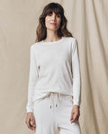 A person stands before a beige fabric backdrop, wearing THE GREAT. F25 CORE KNITS The Long Sleeve Slim Tee in Solid True White and matching white drawstring pants—crafted from 100% cotton for comfort and style.