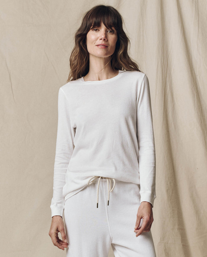 A person stands before a beige fabric backdrop, wearing THE GREAT. F25 CORE KNITS The Long Sleeve Slim Tee in Solid True White and matching white drawstring pants—crafted from 100% cotton for comfort and style.