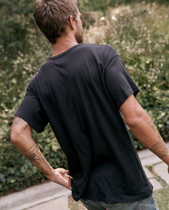 A man with tattoos on his left arm wearing The Men's Boxy Crew in Almost Black by THE GREAT. MAN stands with his back to the camera in a garden setting.