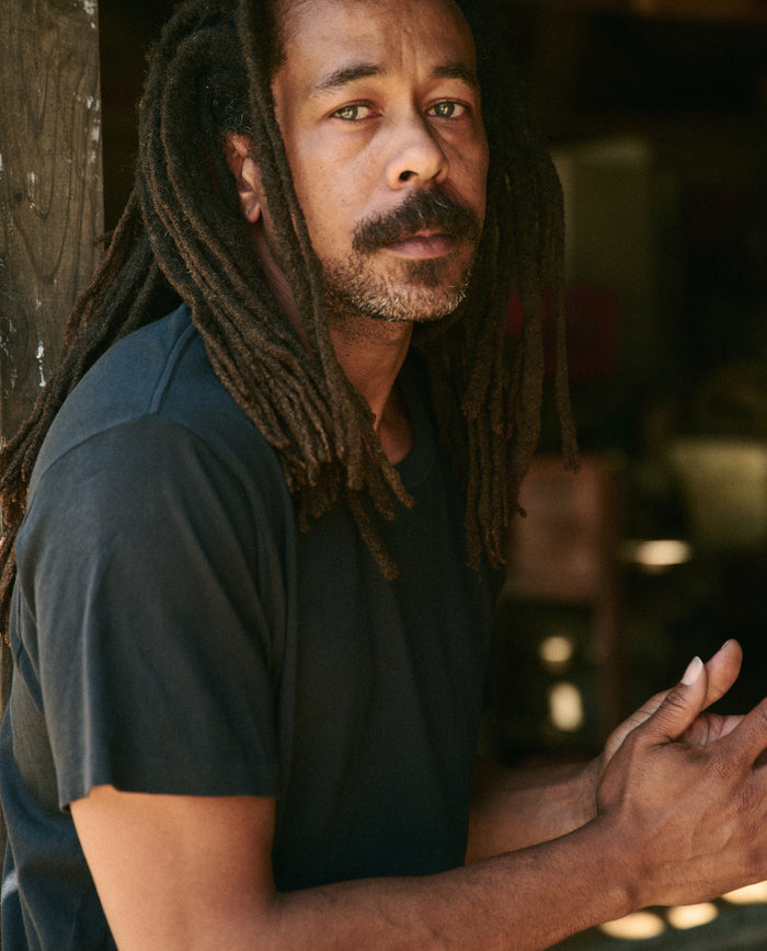 A person with long dreadlocks and a mustache, wearing The Men's Boxy Crew in Almost Black by THE GREAT. MAN, looks at the camera with their hands clasped.