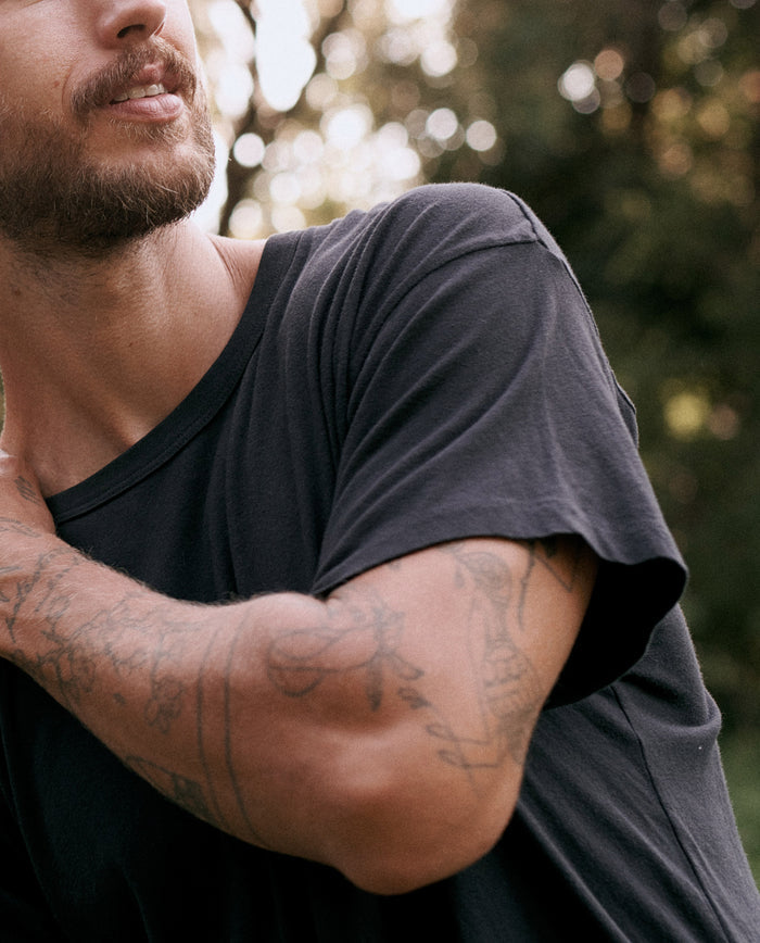 A person with a beard is pictured wearing The Men's Boxy Crew in Almost Black by THE GREAT. MAN, a classic staple in any wardrobe. Their arm, adorned with several visible tattoos, is slightly raised, and the background is blurred with greenery.