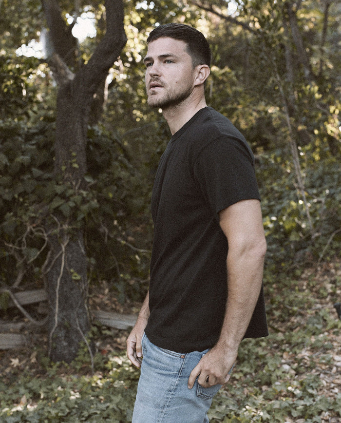 A man, wearing The Men's Boxy Crew in ALMOST BLACK by THE GREAT. MAN and a pair of jeans, stands looking off to the side in a wooded area.