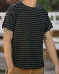 A person wearing The Men's Boxy Crew in Black and Charcoal Stripe by THE GREAT. SU22 MENS, with tattoos on both arms, is standing outdoors with a blurred background. Made in the U.S.A.