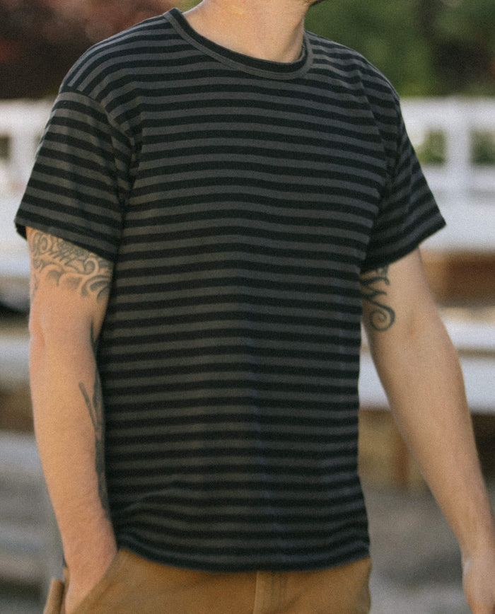 A person wearing The Men's Boxy Crew in Black and Charcoal Stripe by THE GREAT. SU22 MENS, with tattoos on both arms, is standing outdoors with a blurred background. Made in the U.S.A.