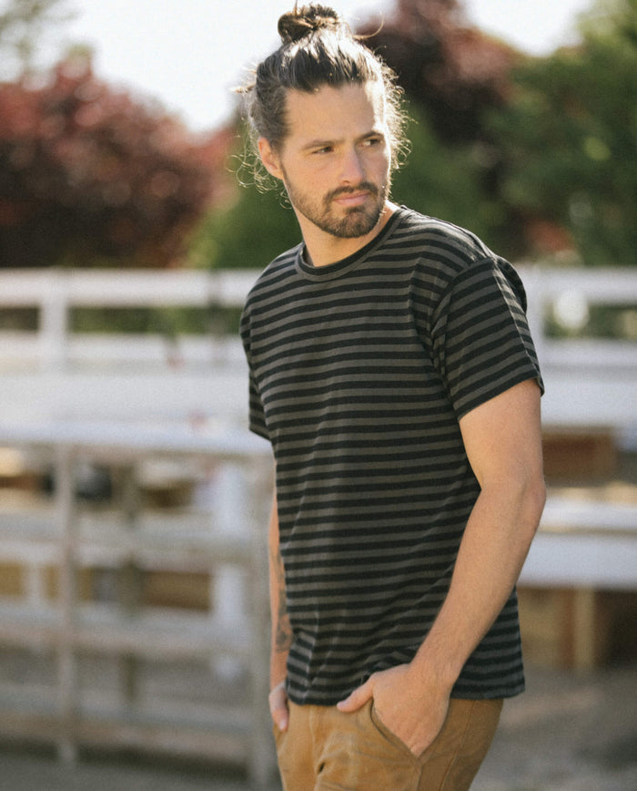 A man with long hair tied up in a bun, wearing "The Men's Boxy Crew" in Black and Charcoal Stripe by THE GREAT. SU22 MENS, and brown pants, stands outdoors with his hands in his pockets.