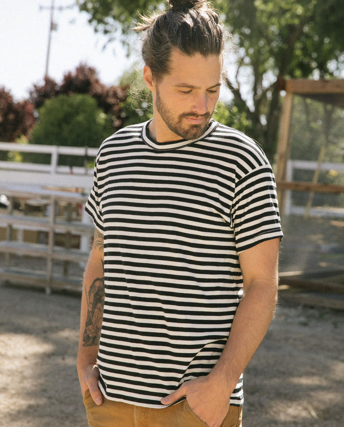 A man with a beard and a man bun, wearing The Men's Boxy Crew in black and cream stripe by THE GREAT. SU22 MENS and brown pants, stands outdoors with his hands in his pockets, looking down.