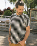 A man with a beard and a man bun, wearing The Men's Boxy Crew in black and cream stripe by THE GREAT. SU22 MENS and brown pants, stands outdoors with his hands in his pockets, looking down.