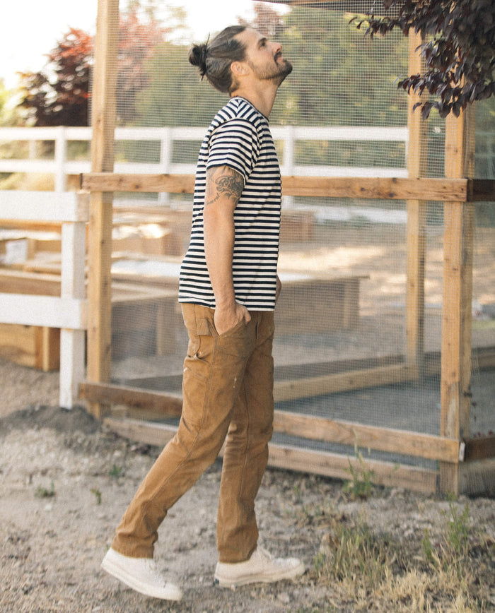 A man in a classic staple, The Men's Boxy Crew in Black and Cream Stripe by THE GREAT. SU22 MENS, paired with brown pants and white sneakers, stands outside looking up with his hands in his pockets. His casual wardrobe choice is complemented by the wooden fence and greenery in the background.