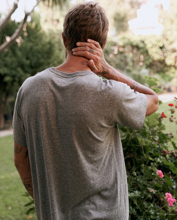 A person with short hair wearing The Men's Boxy Crew in Heather Grey by THE GREAT. MAN stands outdoors, facing away, holding the back of their neck with one hand. A garden with green shrubs and blooming flowers is in the background.