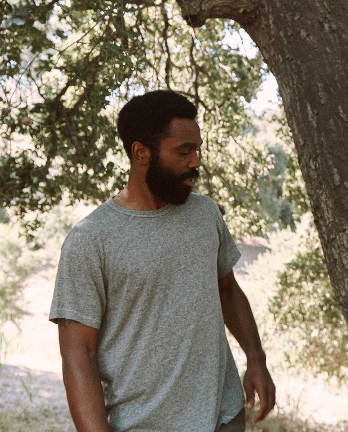 A bearded man wearing The Men's Boxy Crew in heather grey by THE GREAT. MAN stands next to a tree outdoors.