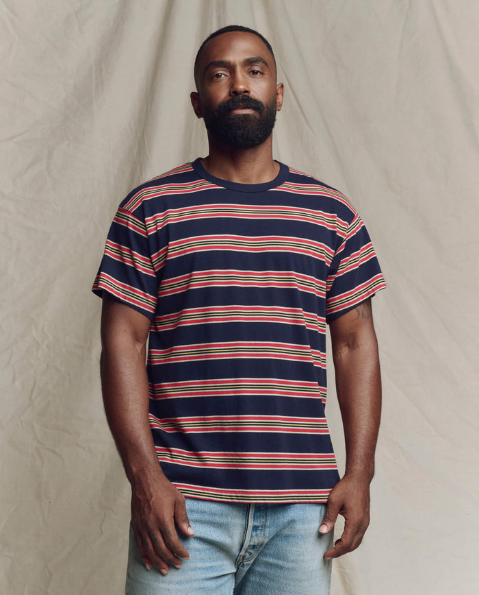 A bearded man stands against a neutral background, wearing THE GREAT. SP26 SEASONAL CORE The Men's Boxy Crew. -- Scout Stripe and light blue jeans.