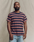 A bearded man stands against a neutral background, wearing THE GREAT. SP26 SEASONAL CORE The Men's Boxy Crew. -- Scout Stripe and light blue jeans.