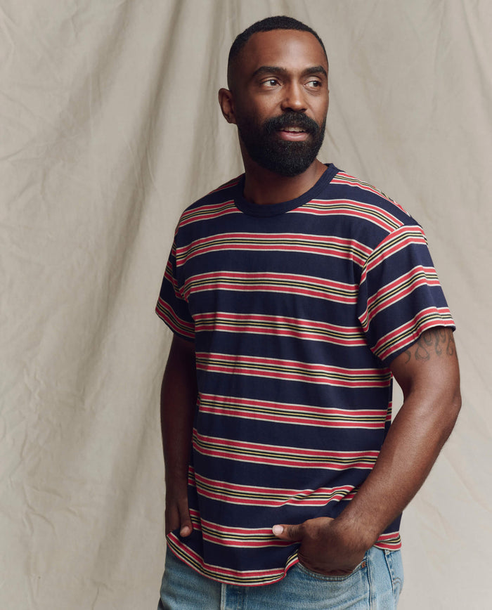 A bearded man wears THE GREAT.'s SP26 SEASONAL CORE The Men's Boxy Crew. -- Scout Stripe tee in navy, red, and white yarn-dye stripes with light blue jeans, standing with hands in pockets against a beige fabric backdrop.